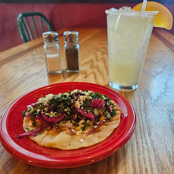 A Mexican tostada with purple cabbage, cilantro, and cheese, next to a margarita on a wooden table.