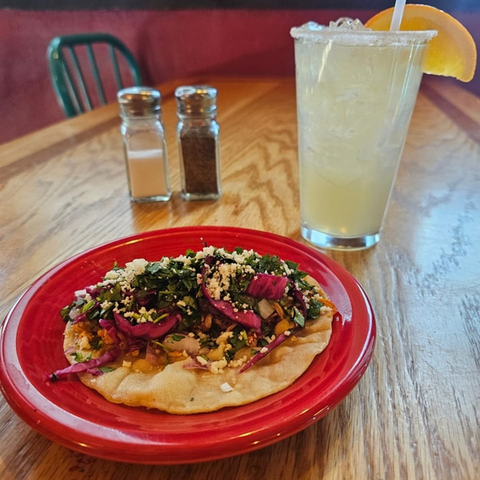 A Mexican tostada with purple cabbage, cilantro, and cheese, next to a margarita on a wooden table.
