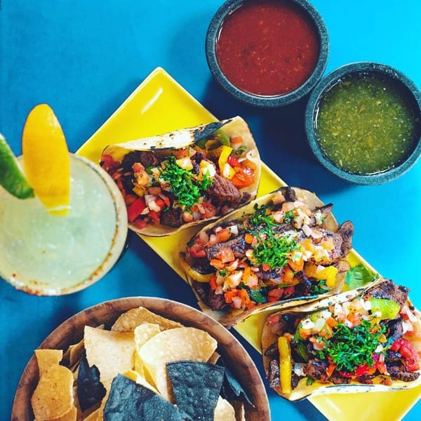 Three steak tacos with pico de gallo, a margarita, chips, red and green salsa on a blue table.