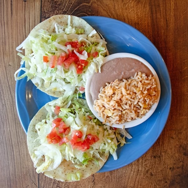 Two Mexican tacos with lettuce and tomatoes, served with rice and refried beans on a blue plate.